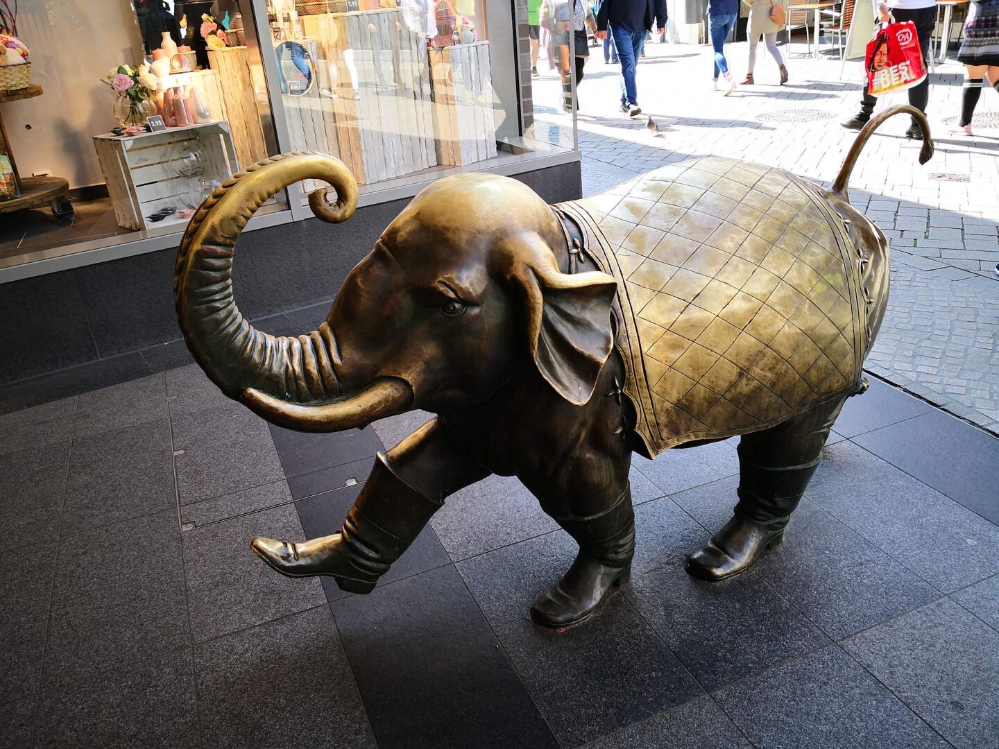 Zu sehen ist ein Foto, das einen kleinen Elefanten aus Bronze zeigt, der mit einer Decke (ebenfalls Bronze) und Stiefeln an seinen Beinen in ein Geschäft in der Oldenburger Innenstadt läuft.
Die Statue ist im - äusseren, also noch auf der Seite der Fußgängerzone und nicht IM Ladengeschäft - montiert. Kopf, Rüssel und Decke glänzen goldfarben, was auf viele Berührungen hinweist, Beine und Bauch sind eher olivdunkel.