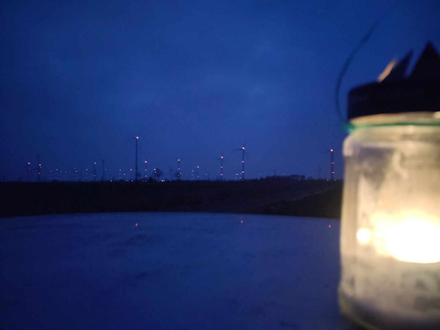 Das Foto zeigt in einiger Entfernung eine Reihe von Windenergieanlagen, die vor dem spät dämmernden Abendhimmel mit Ihren „Hinderniskennzeichnungen“ / Flugbeleuchtungen erstrahlen.

Im Vordergrund steht am rechten Bildrand eine brennende Kerze in einem durchsichtigen Gefäss mit Deckel.