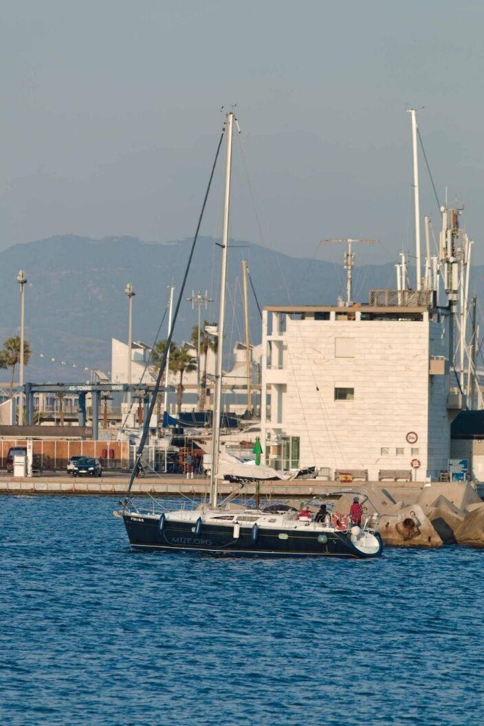 Ein Segelschiff fährt, ohne Segel und mit Motor, in den Hafen ein. Im Hintergrund sind die Hafenanlagen und ein Berg zu sehen./ A sailing ship enters the harbor without sails and with a motor. The harbor facilities and a mountain can be seen in the background.
