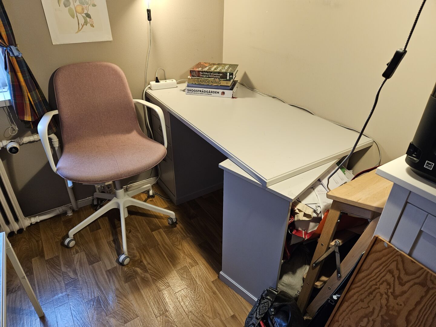 My current desk, a tiny grey IKEA unit with a loose desktop resting on a drawer and a shelving unit. The shelving unit stands askew since I move around while I work and my legs keep pushing against it.