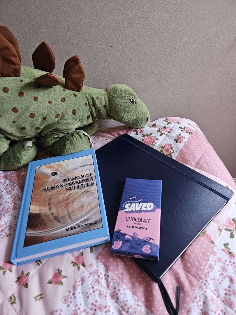 A plush stegosaurus guarding my blue project notebook, the book "Design of Human-Powered Vehicles" by Mark Archibald, and a bar of "Saved Chocolate Mixed by Motatos".