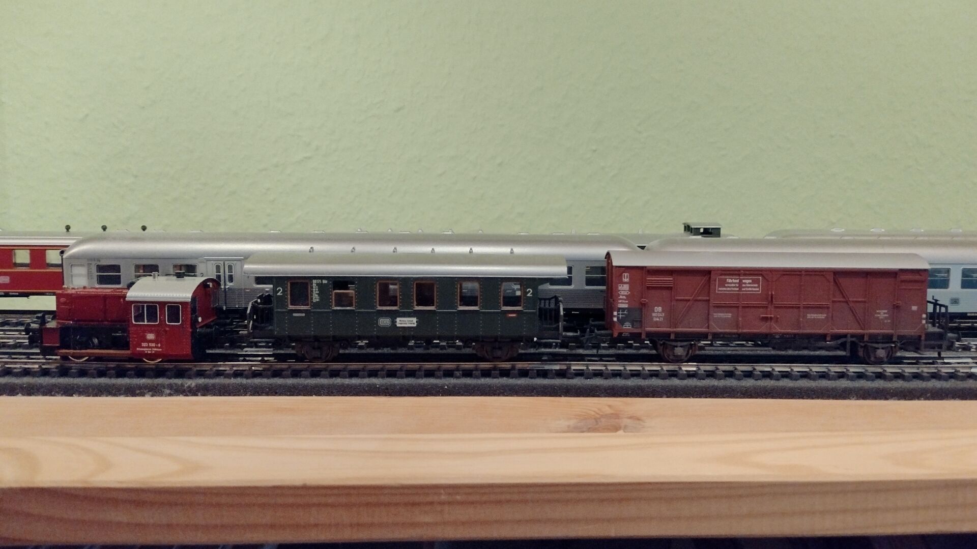 H0-Modell eines Güterzugs mit Personenbeförderung der DB. Eine rote Kleinlok der Bauart Köf II, ein grüner Einheits-Nebenbahnwagen und ein brauner gedeckter Fährboot-Güterwagen auf Gleisen in einem IVAR-Regal. Im Hintergrund Silberling-Nahverkehrswagen und ein roter Steuerwagen.