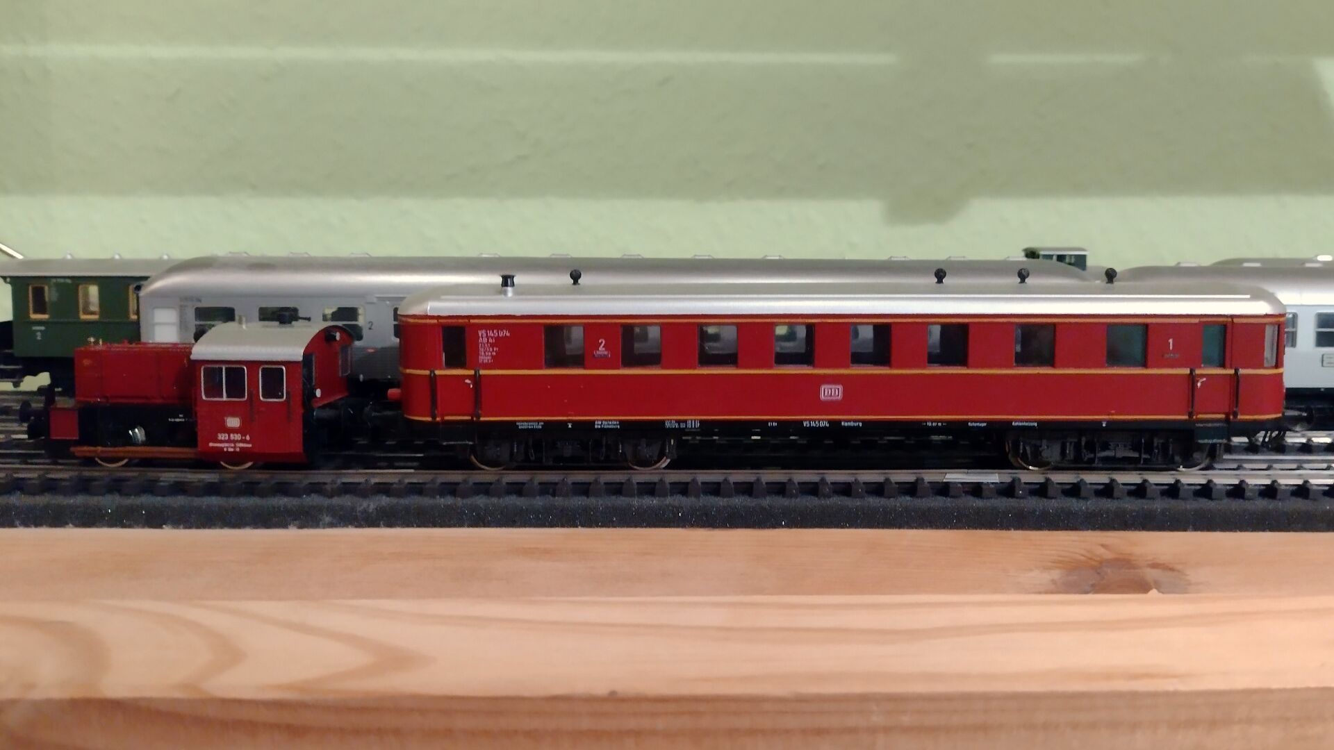 Eine rote Kleinlok der Baureihe Köf II (beschriftet als Baureihe 323) der DB zieht einen roten vierachsigen Beiwagen der Reichsbahn-Bauart der DB. Im Hintergrund Silberlinge und grüne zweiachsige Personenwagen. H0-Modelle in einem IVAR-Regal