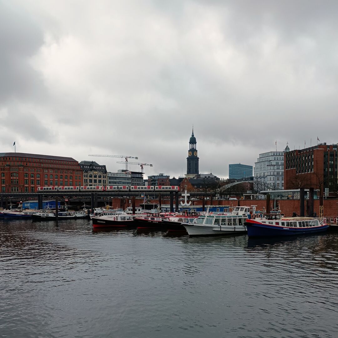 Blick vom Kehrwieder in der Hamburger Speicherstadt in Richtung Michel. Im Vordergrund die ruhige, graue Wasserfläche eines Fleets, dahinter Barkassen und die Flussschifferkirche, ein Hochbahn-Viadukt mit einem rot-weißen Hochbahnzug, Gebäude, zwei Baukräne und der Turm der Michaeliskirche. Der Himmel ist bedeckt und grau.