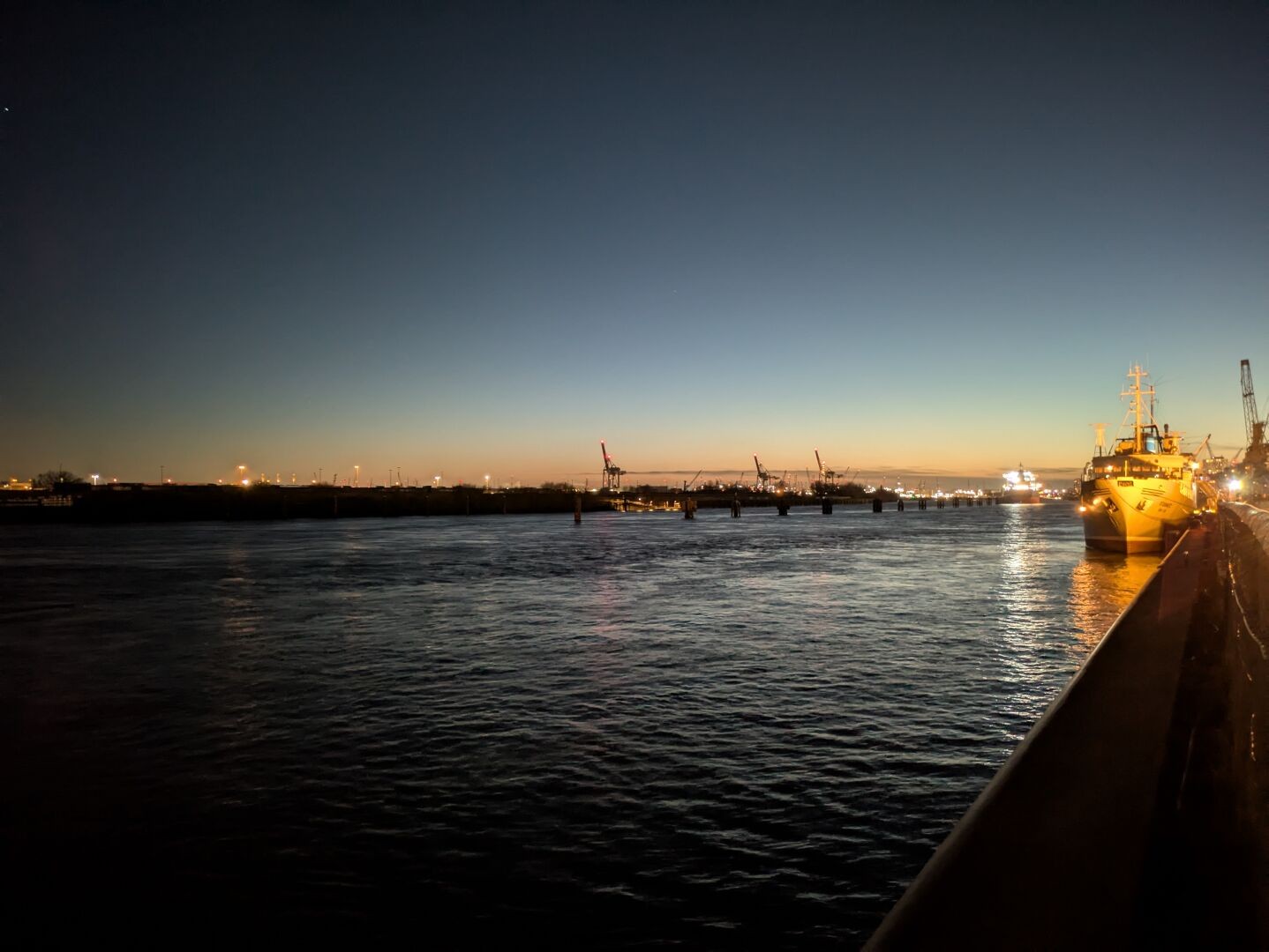 Blick von der Kaimauer auf die MS Stubnitz, die an ihrem Ankerplatz liegt (rechts im Bild). Es ist klarer Himmer, die Sonne geht am Horizont unter. Links vom Schiff ist das gegenüberliegende Elbufer mit einigen Hafenkränen zu sehen.