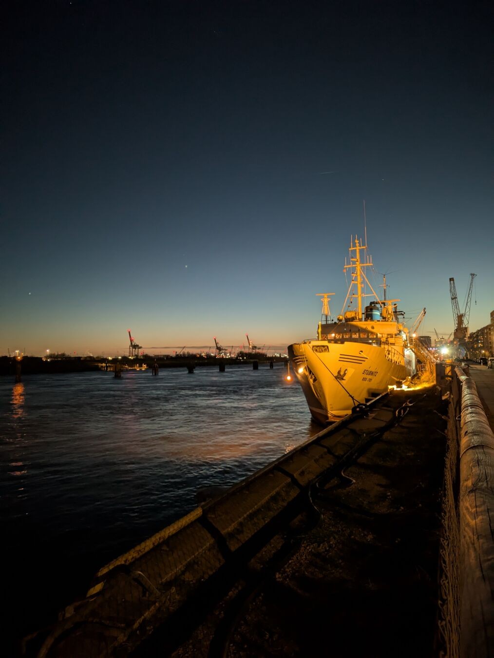 Blick von der Kaimauer auf die MS Stubnitz, die an ihrem Ankerplatz liegt. Es ist klarer Himmer, die Sonne geht am Horizont unter. Im Hintergrund des Schiffs ist das gegenüberliegende Elbufer mit einigen Hafenkränen zu sehen.