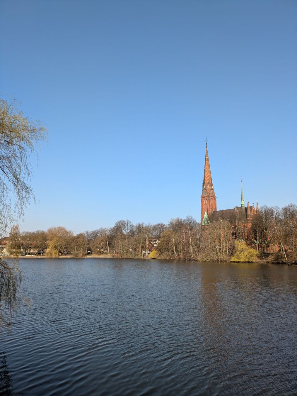 Blick über den Kuhmühlenteich Richtung gegenüberliegendes Ufer. Dort stehen viele Bäume und im Hintergrund ist die Kirche St. Gertrud zu sehen. Es ist strahlend blauer Himmel ohne eine einzige Wolke.