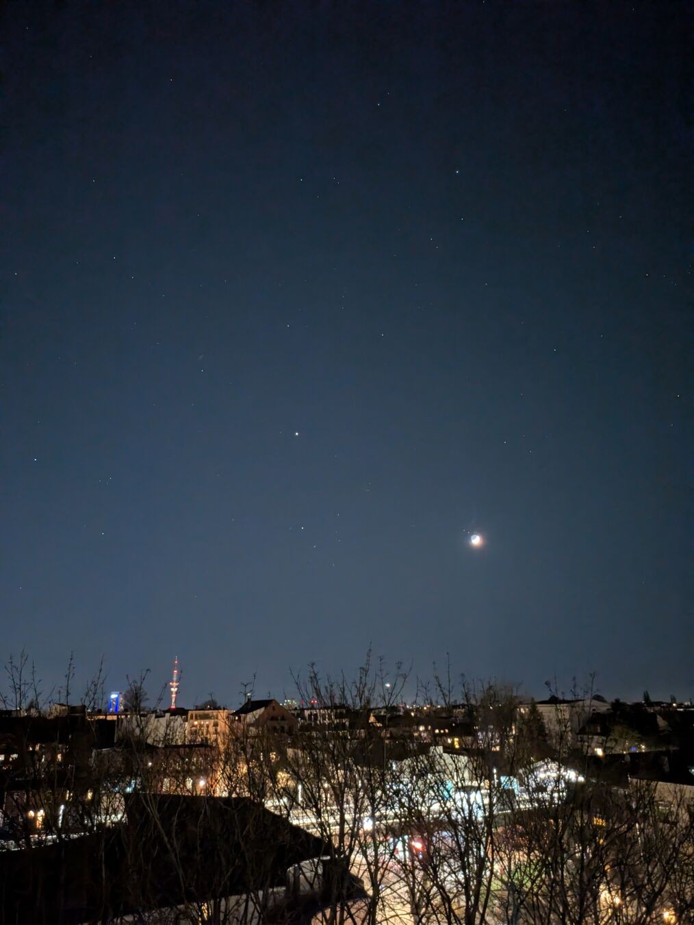 Blick in den klaren Abend-/Nachthimmel, der voller Sterne ist und einen hell leuchtenden zunehmenden Mond zeigt. Im unteren Bildviertel ist ein Teil der noch stark beleuchteten Stadt zu sehen, der Blick schweift über die Häuserdächer.