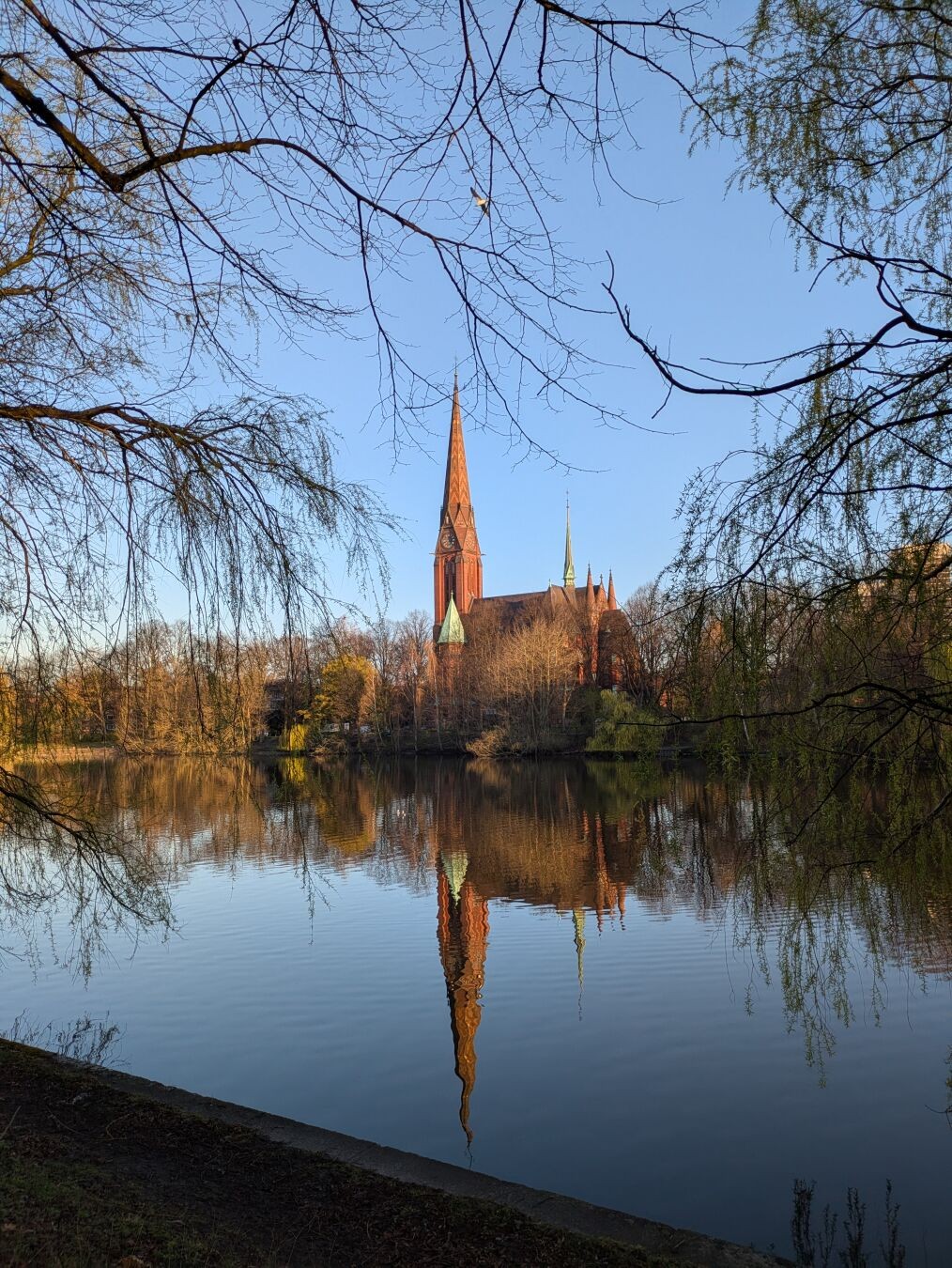 Blick über den Kuhmühlenteich auf die am gegenüberliegenden Ufer befindliche St. Gertrud-Kirche. Am linken und rechten Bildrand sind jeweils herunterragende Äste am Wegesrand stehender Bäume zu sehen.