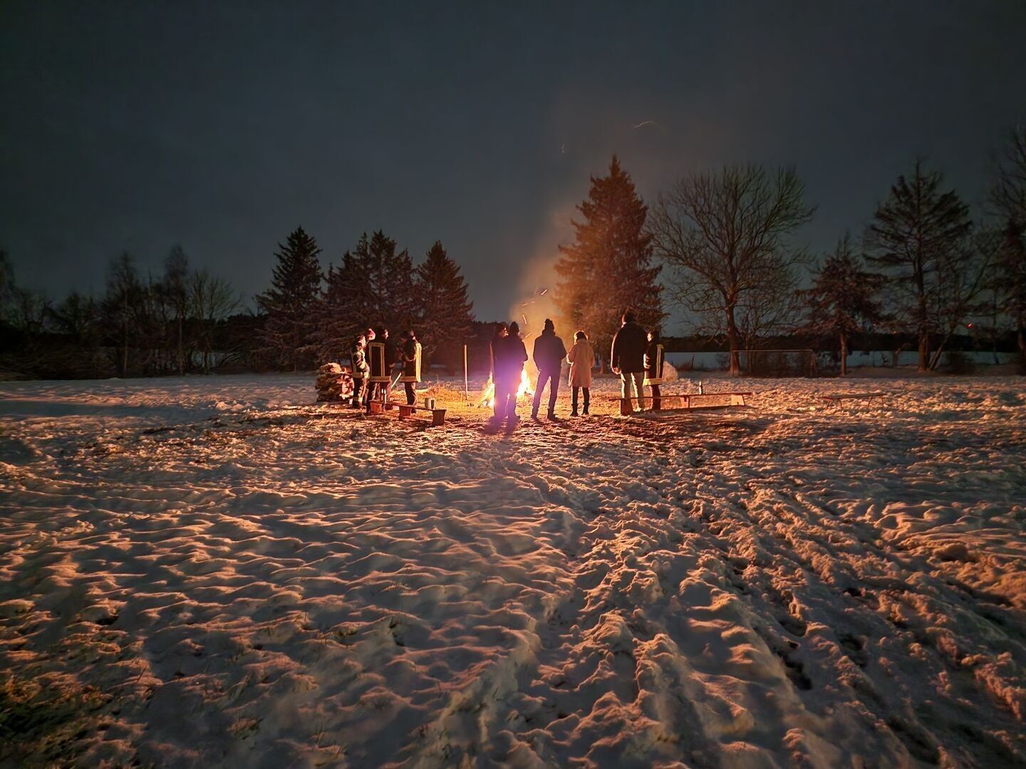 People standing around an camp fire at a field covered with snow in the night.