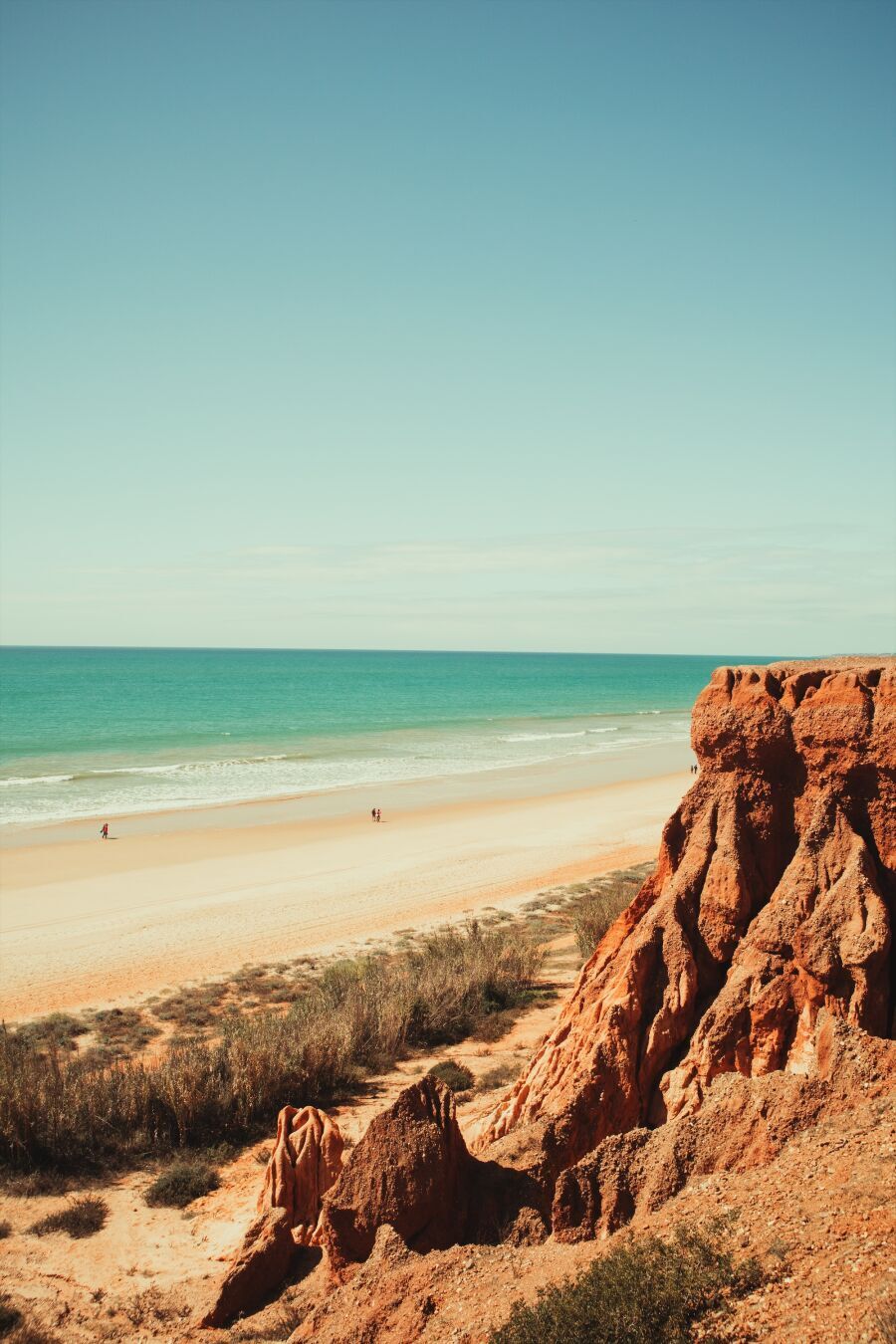 A scenic coastal landscape featuring rugged reddish-brown cliffs in the foreground and on the right, sloping down to a sandy beach that stretches into the distance. The beach meets clear turquoise waves on the left rolling gently onto the shore. Sparse vegetation grows on the cliffs and near the beach. A few people are walking along the shoreline under a vast blue sky with minimal clouds. In the distance, a small town can be seen.