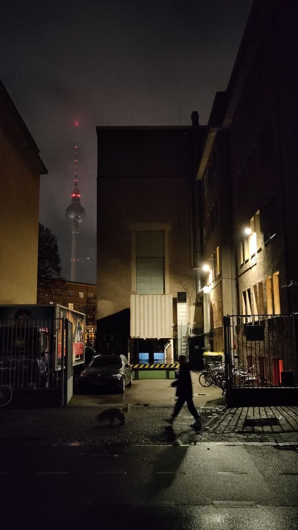 Walking the dog at night, passing by an open Berlin backyard with the TV tower looming in the background in a gap between two buildings