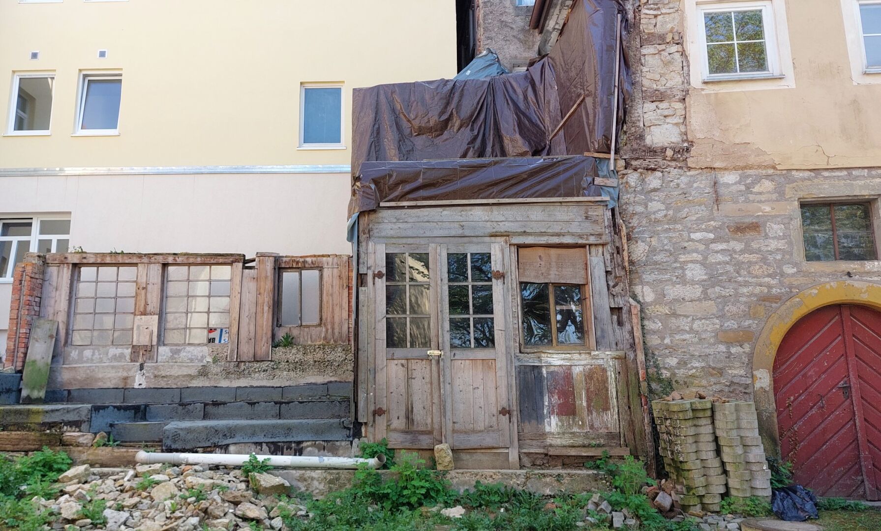 Foto eines zu großen Teilen abgerissenen Hauses, aus Backsteinen und Naturstein gemauert mit großen Holzanteilen an Türen und Fenstern. Vom abgerissenen Hausteil steht noch eine Rückwand mit einer Fensterfrlnt und der Eingangsbereich mit ungestrichener zweiflügleliger Holzeingangstür und einem kleinen Fenster direkt daneben.
Direkt hinter dem abgerissenen Hausteil liegt die hell gestrichene Wand eines Neubaus.