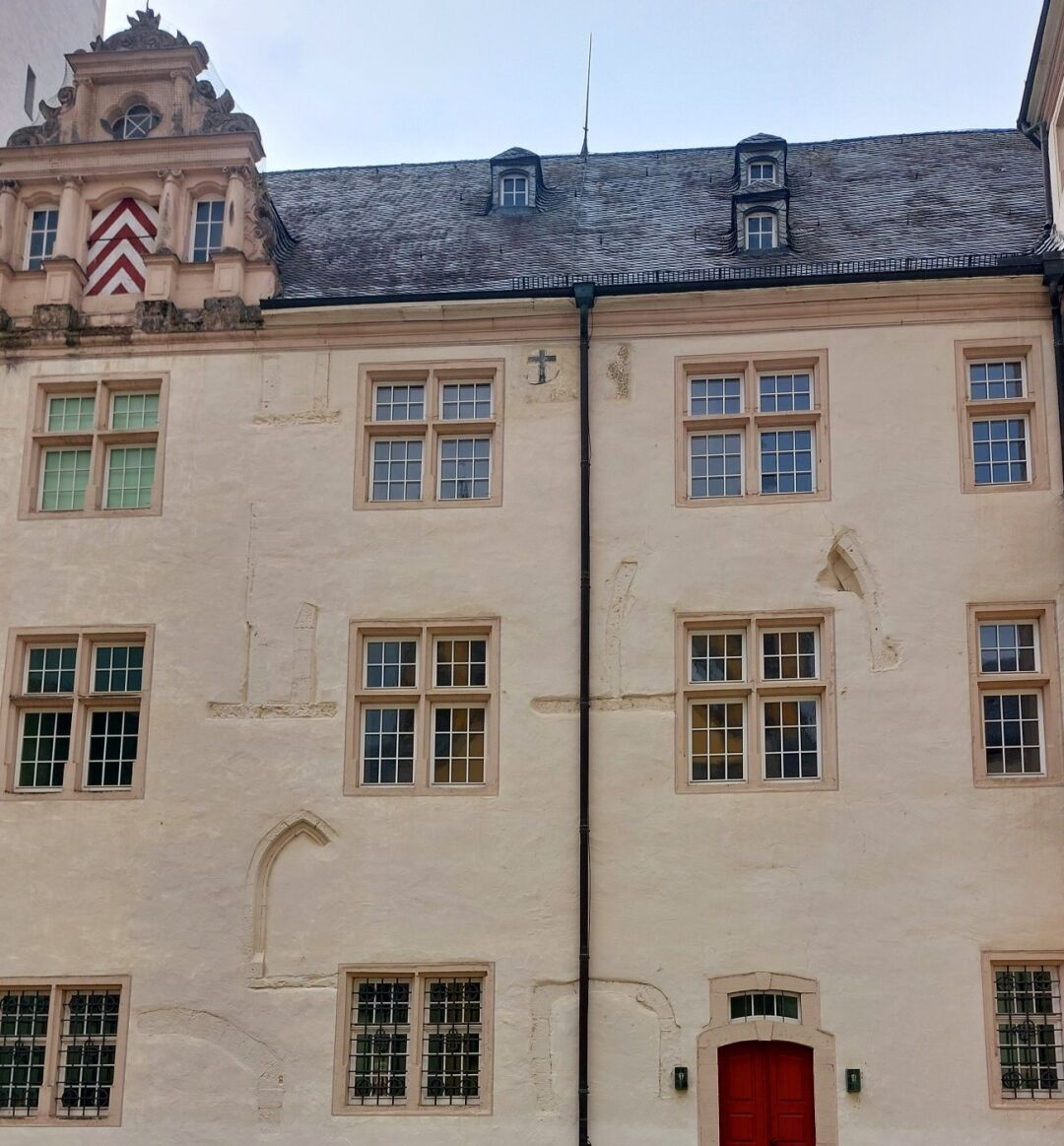 Foto einer beige gestrichenen Fassade eines dreistöckigen Gebäudes mit zusätzlichen Geschossen im Dach.
An der Fassade sind regelmäßig rechteckige Fenster mit jeweils 2 geteilten Fensterflügeln angeordnet. Dazwischen sind im Mauerwerk nicht vollständig verputzt die Umrisse älterer gemauerter Fensterrahmen auf unterschiedlichen Höhe  zu sehenn mit unterschiedlichen Höhen und verschiedenen Bogenformen.