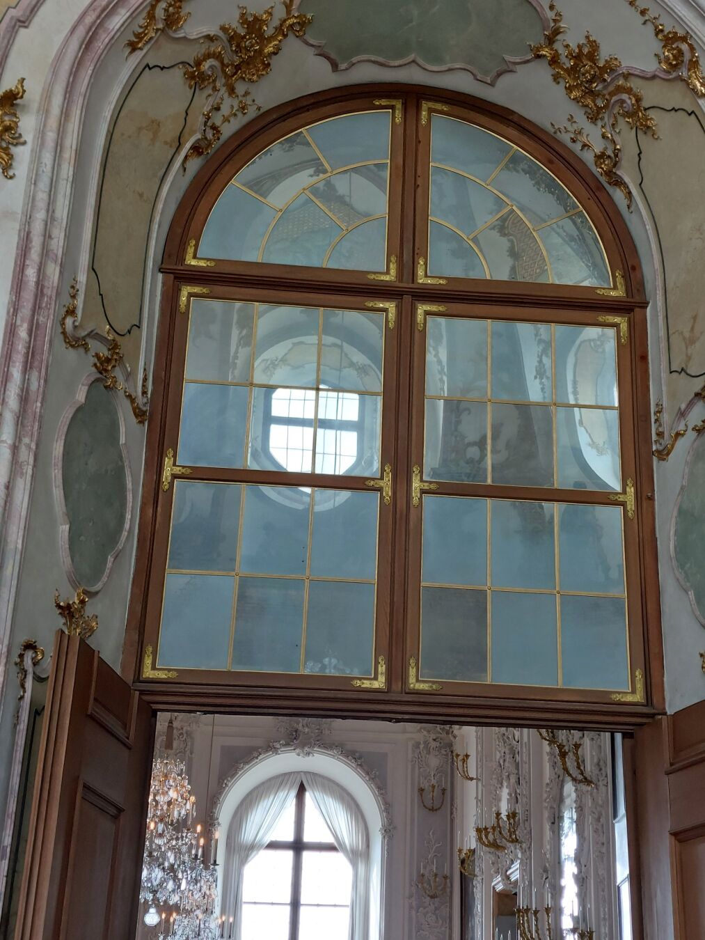 Foto eines verzierten barocken Holzrahmenfensters aus mehreren Segmenten mit halbrunden Abschluss. Anstelle der Glasscheiben sind unterschiedlich angelaufene Spiegel in die Fenstersegmente eingesetzt.
In den Spiegeln ist die Spiegelung eines  Deckengwölbes mit einem hohen Lichtfenster zu sehen.
Unter dem Spiegel ist eine geöffnete Tür, dahinter liegt ein heller Saal mit einem Fenster gegenüber der Tür.
