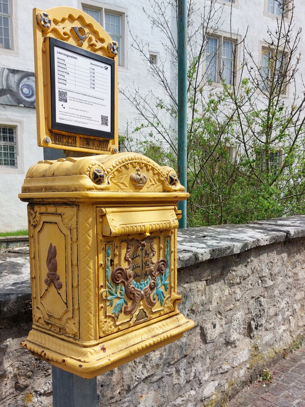 Farbiges Foto eines altmodischen Briefkasten vor einer niedrigen Steinmauer. Der Briefkasten etwas blassgelb mit farbigen Akzenten gestrichen und aufändig verziert geformt. Die Verzierungen bestehen aus verspielten Ranken und Ornamentem sowie einem Stadtwappen und einer Krone oberhalb des Einwurfs.
Der Briefkasten ist an einem modernen verzinkten Vierkantrohr angebracht, Oberhalb ist eine ebenfalls verzierte blassgelbe Blechtafel angebracht, auf der ein moderner Zettel mit den Leerungszeiten angebracht ist.