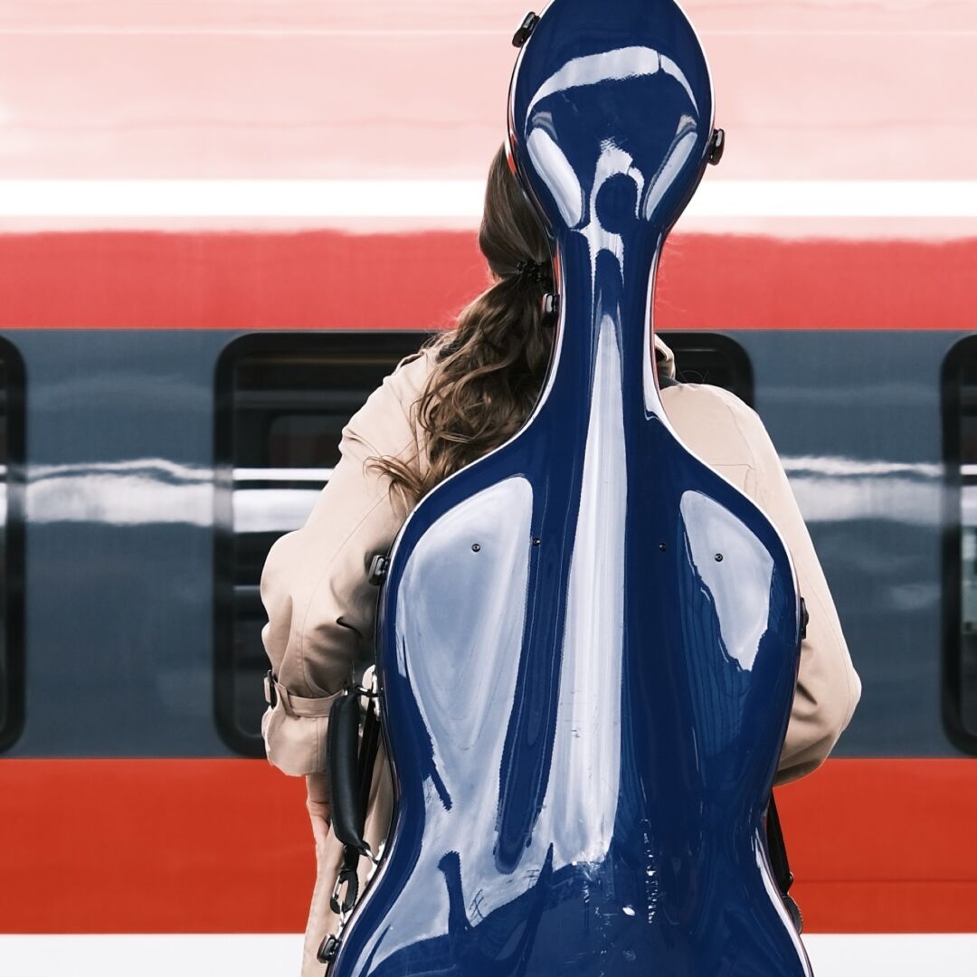 Vor einem roten Zug steht eine Person mit langen Haaren und einem glänzenden blauen Cellokasten auf dem Rücken.

A person with long hair and a shiny blue cello case on their back is standing in front of a red train.