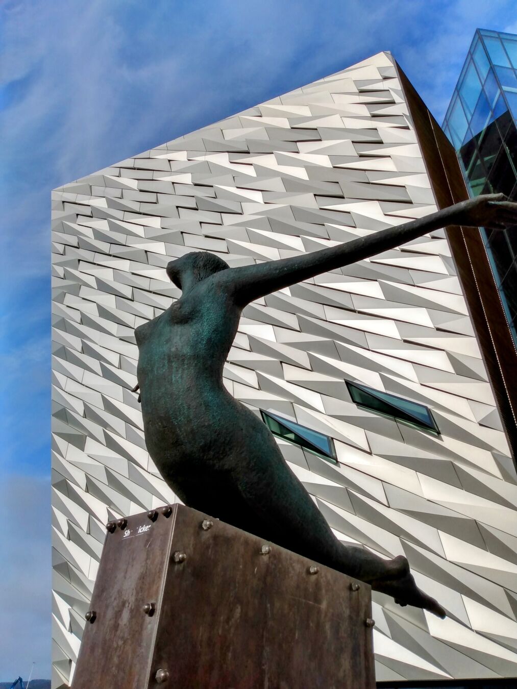 A shiny, silvery front of a modern building under a blue sky, reminiscent of a school of fish or waves. In front of it, a female metal sculpture stretches upwards on a stone plinth with outstretched arms, like the figures on the bow of old sailing ships.
Eine silbrig glänzende Front eines modernen Gebäudes unter einem blauen Himmel, die an einen Fischschwarm oder Wellen erinnern könnte. Davor streckt sich auf einem Steinsockel eine weibliche Metallskulptur mit ausgebreiteten Armen nach vorne oben, so wie die Figuren auf dem Bug alter Segelschiffe. A shiny, silvery front of a modern building under a blue sky, reminiscent of a school of fish or waves. In front of it, a female metal sculpture stretches upwards on a stone plinth with outstretched arms, like the figures on the bow of old sailing ships.
Eine silbrig glänzende Front eines modernen Gebäudes unter einem blauen Himmel, die an einen Fischschwarm oder Wellen erinnern könnte. Davor streckt sich auf einem Steinsockel eine weibliche Metallskulptur mit ausgebreiteten Armen nach vorne oben, so wie die Figuren auf dem Bug alter Segelschiffe.