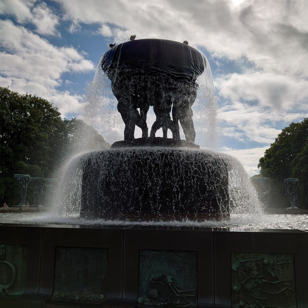 A three tiered fountain, about 7-8 metres tall. The bottom tier is a large, rectangular pool. The next tier is a round pedestal. The top tier is a bowl, held up by human statues standing on the pedestal. Water is pouring down. On the top seagulls are resting. In the background there are trees to the left and right, and a blue partly cloudy sky.