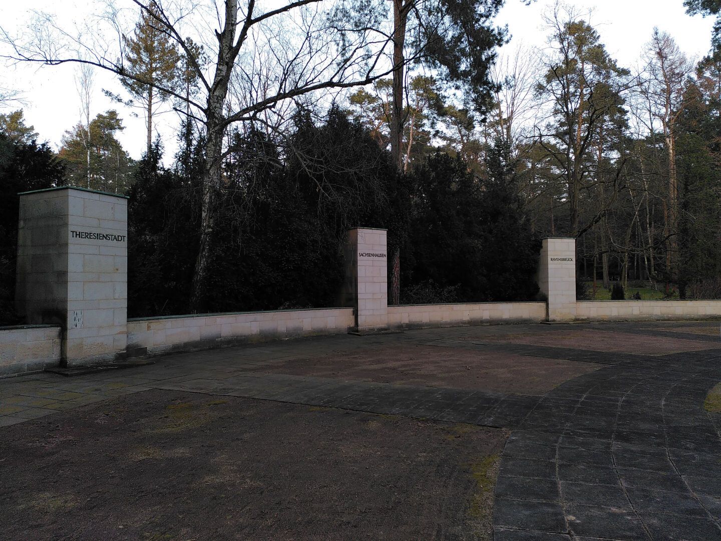 Teil eines Gedenkortes, zu sehen ist ein Ausschnitt einer Kreisfläche, an deren Rand sind drei massive Stelen mit den Beschriftungen "Theresienstadt", "Sachsenhausen", "Ravensbrück"