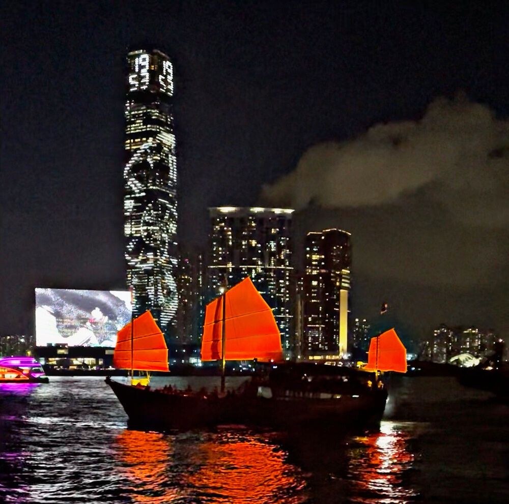 Victoria harbor and a junk ship sailing across the water at night with Hong Kong skyline in the background