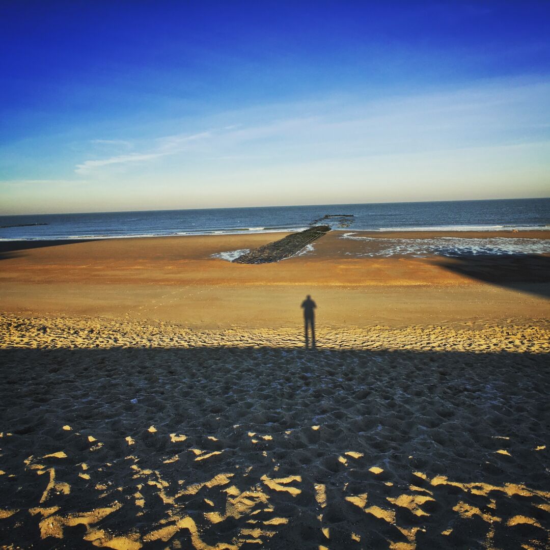 Ein breiter Strand vor dem Meer. Vorn verläuft ein Schatten, der einen großen Teil des Strands einnimmt. In der Mitte ist der Schatten des Fotografen zu erkennen.