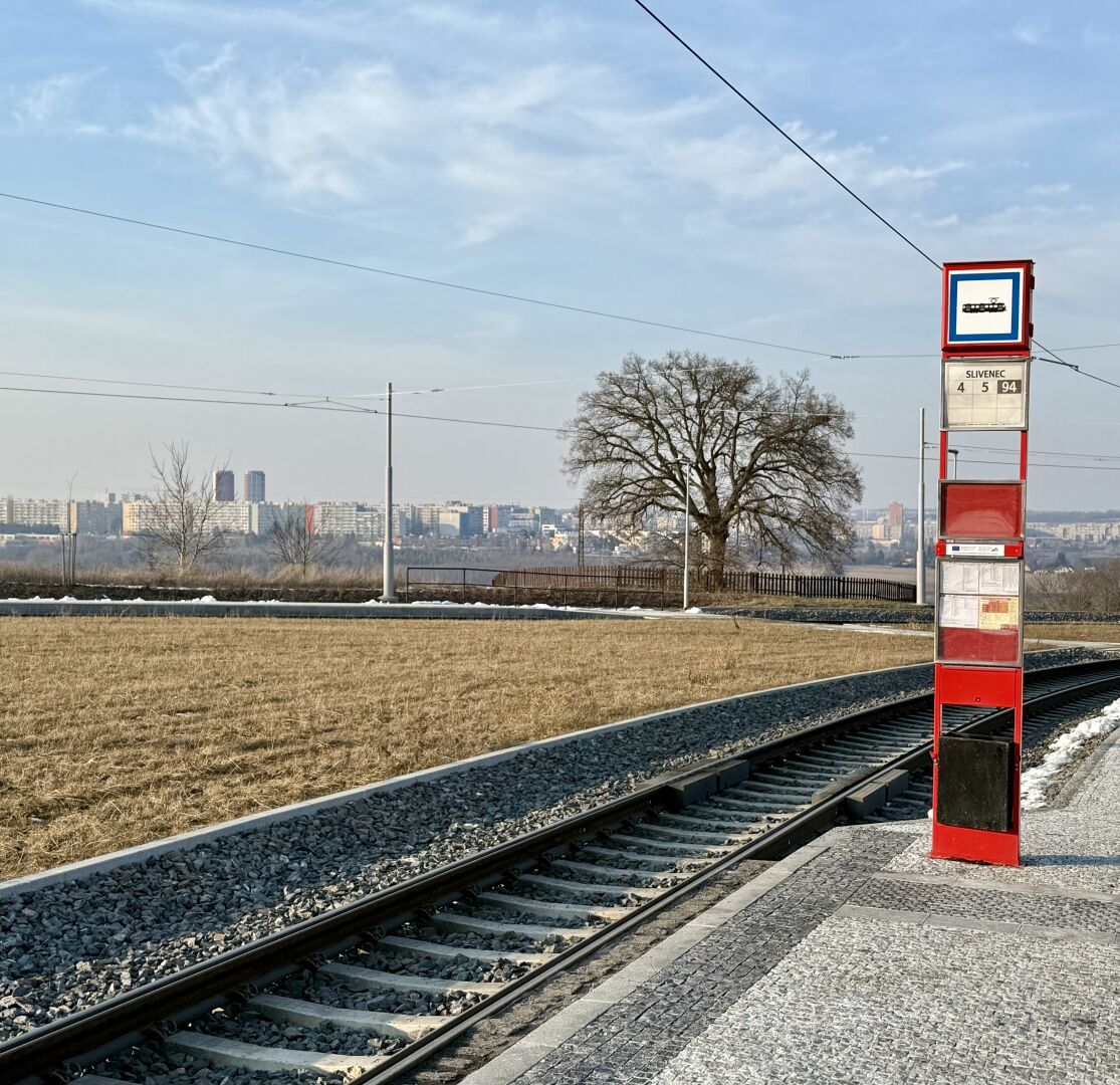 Eine Straßenbahnhaltestelle, erkennbar an der Säule mit Informationen zur hier verkehrenden Linie. Im Hintergrund steht ein einzelner Baum, dahinter ist in einiger Entfernung die Shilouette von Prager Vororten erkennbar.