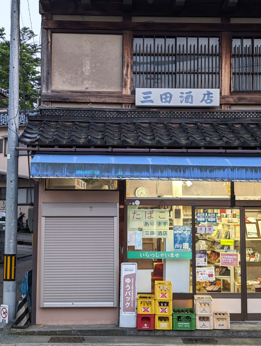 A vintage storefront illuminated from the inside. There are a lot of Japanese signs on the glass window. There are plastic boxes at the front. The second floor of the building is an old-looking Japanese style house.