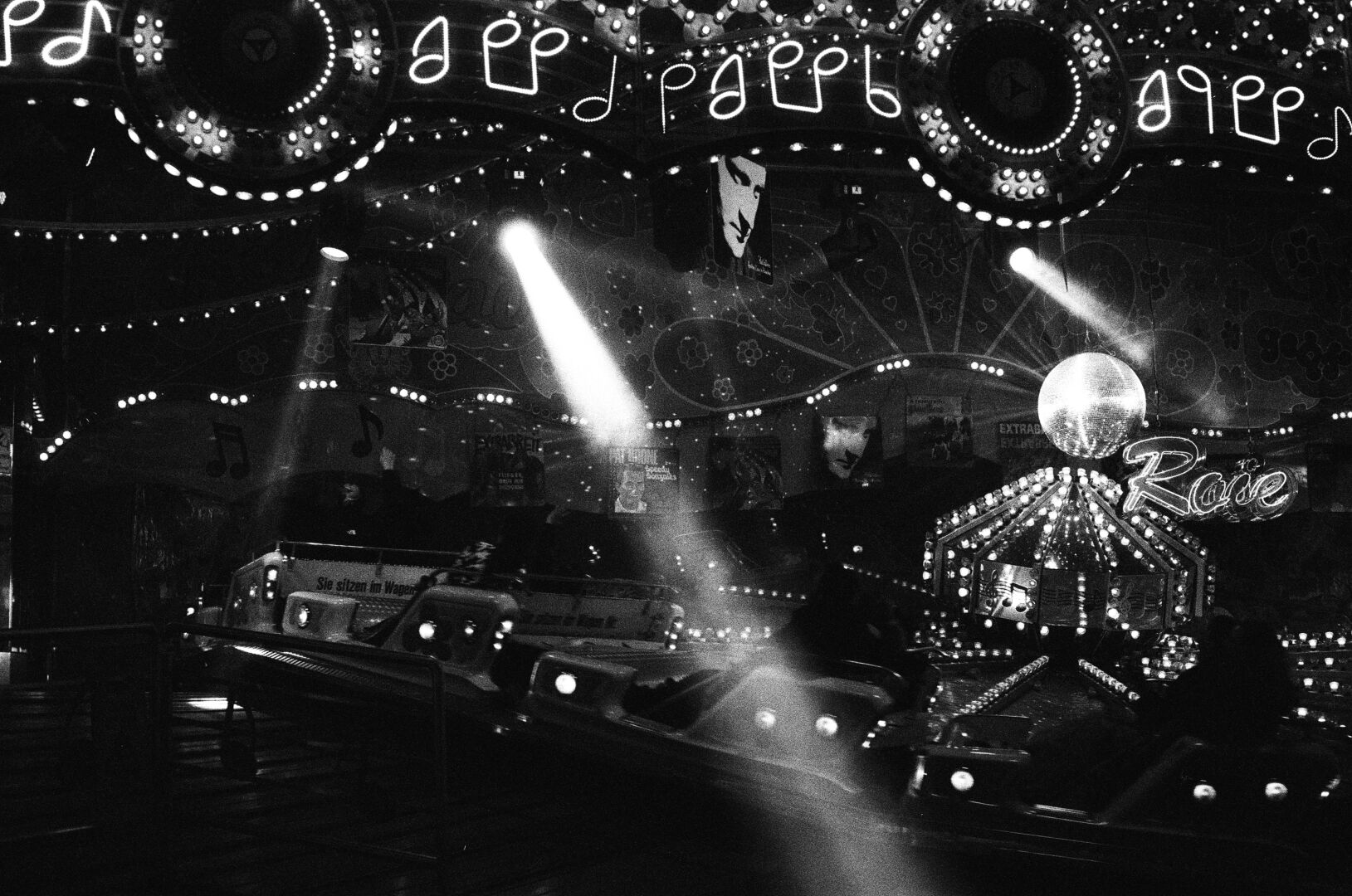 A very high contrast black-and-white photo of a circular fair ride with a disco ball in the middle and lots and lots of lights on the ceiling. Some of the spotlights have their beams visible due to haze
