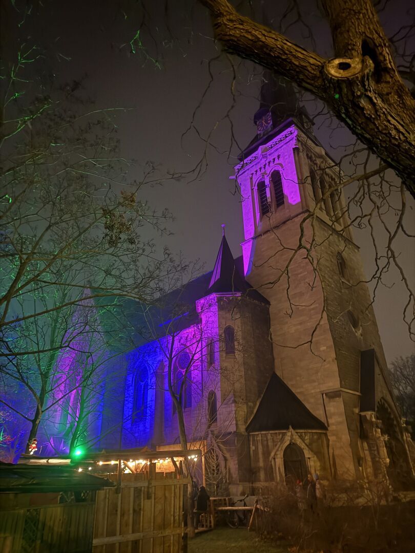 Man sieht im Dunkeln eine Kirche. Rechts im Vordergrund ist der Kirchturm. Die Kirche wird von unten in einem lila Farbton angestrahlt.