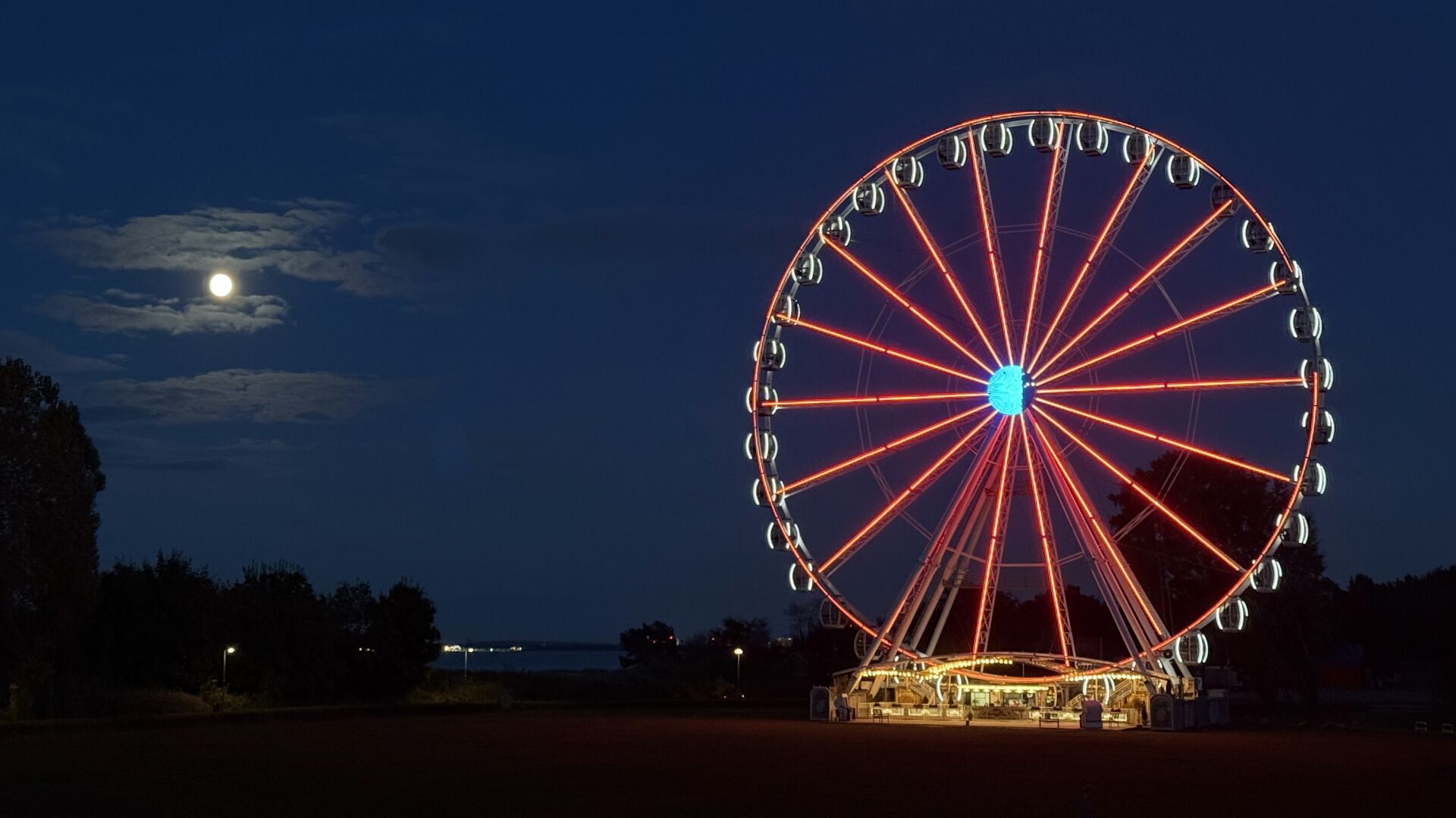 Nachtaufnahme eines rot beleuchteten Riesenrads im rechten Teil. Links sieht man den Mond hinter Wolken.