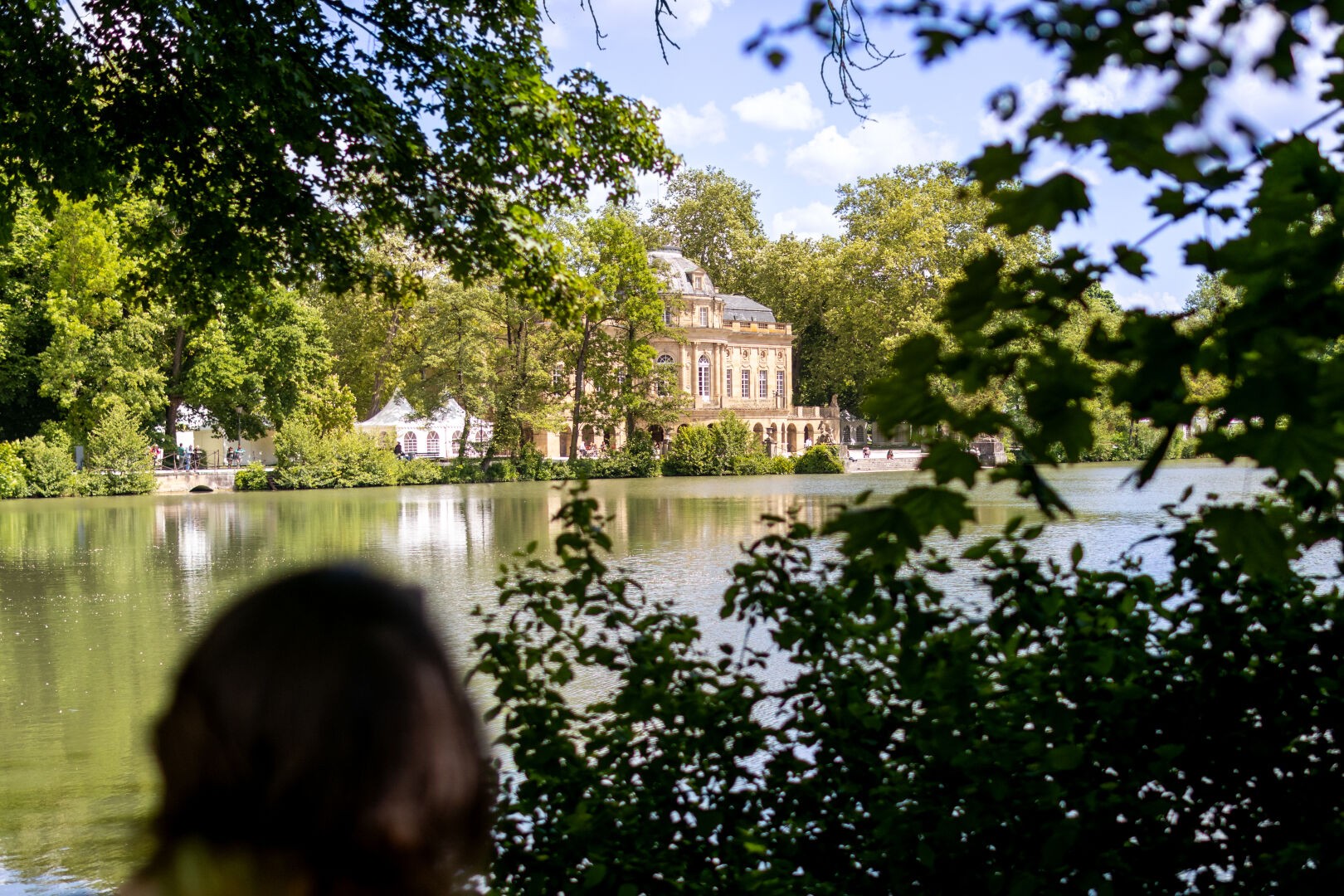 Ein Foto eines Schlosses an einem See. Das Schloss ist durch eine Lücke in der Vegetation fotografiert, das Bild ist an den Seiten von grünen Pflanzen eingerahmt. Im Hintergrund ist der blaue Himmel mit einigen weißen Wolken zu sehen. Vorne links ist der Kopf einer Person in der Unschärfe zu erahnen.