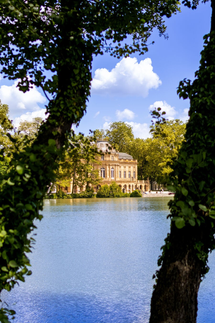 Ein Foto eines Schlosses an einem See. Das Bild ist links und rechts von Bäumen im Vordergrund eingerahmt. Im Hintergrund ist der blaue Himmel mit einigen weißen Wolken zu sehen.
