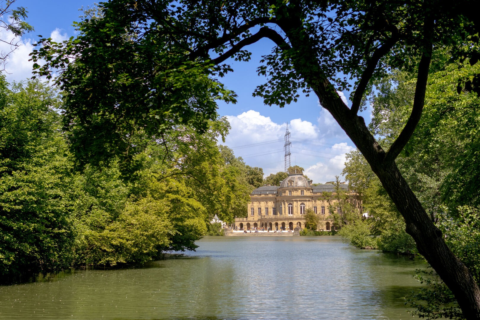 ein Foto von einem Schloss an einem See. Der See nimmt die untere Hälfte des Bildes ein. Schloss und See sind von grünen Pflanzen eingerahmt, im Hintergrund ist der blaue Himmel mit einigen Wolken zu sehen. Im Vordergrund ragt ein Baum schräg in das Bild.