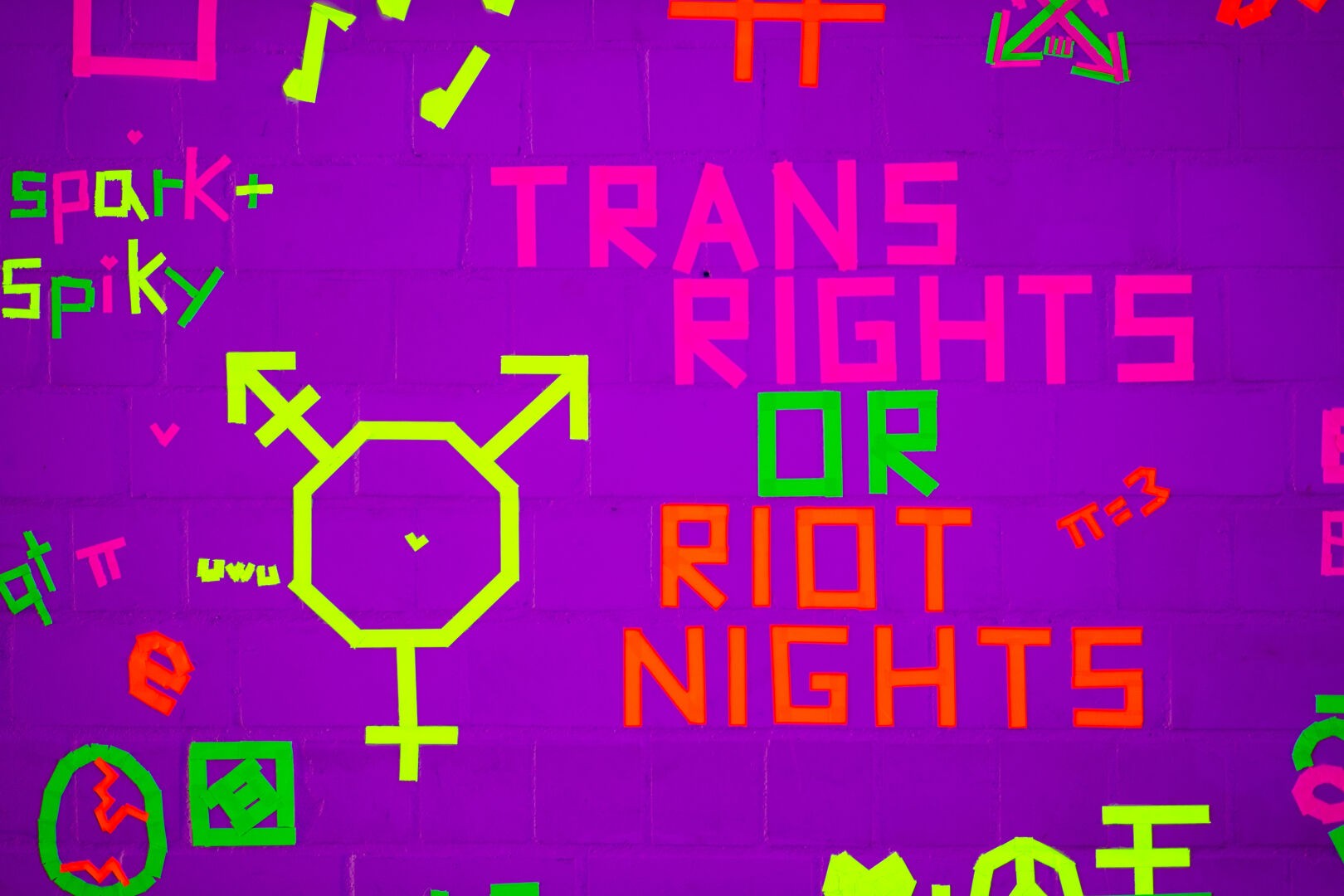 Schwarzlichtkunst. Mit Neon-Tape sinf verschiedene Motive und der Text "TRANS RIGHTS OR RIOT NIGHTS" auf eine gemauerte Wand geklebt.