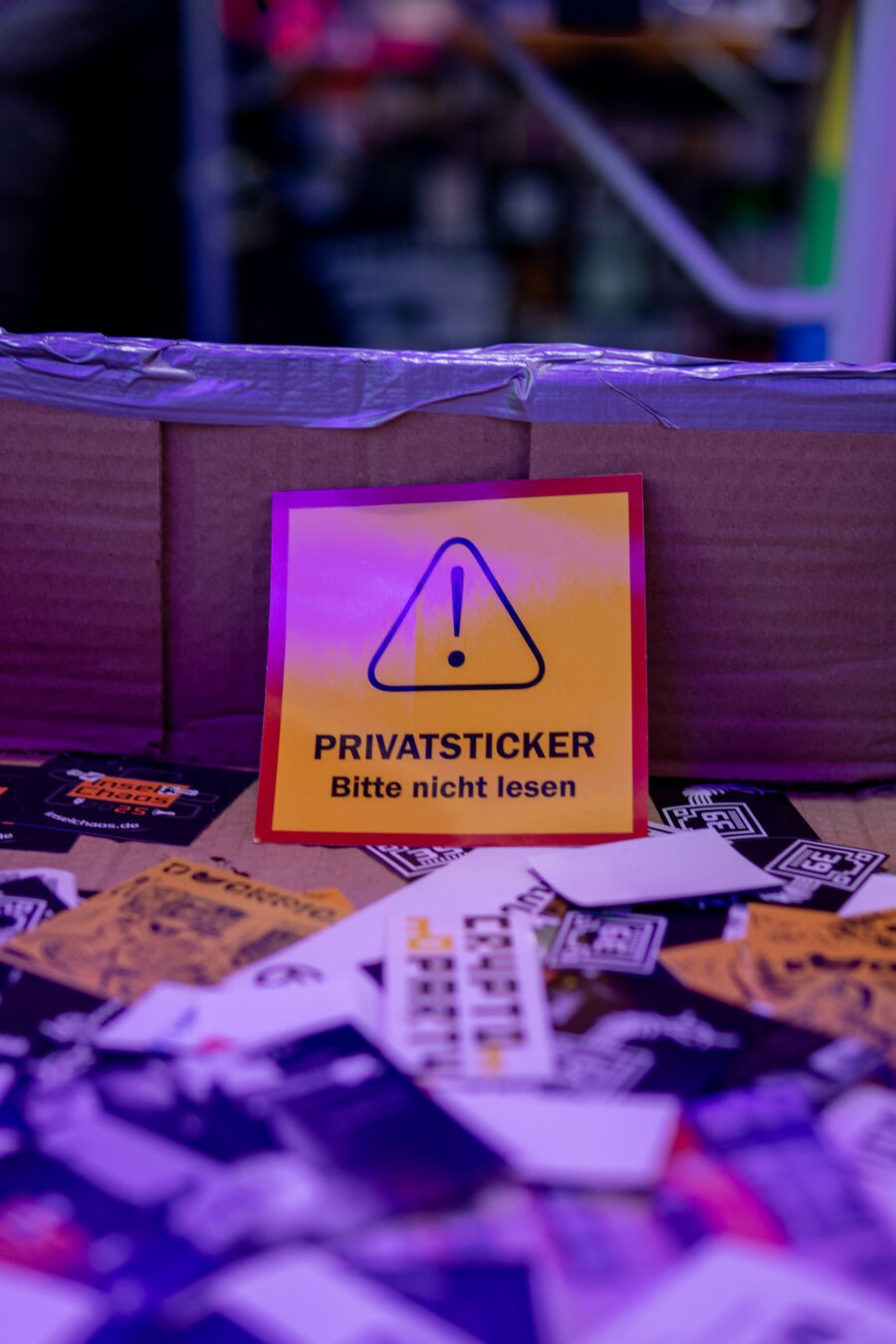 Ein Pappkiste voller unterschiedlicher Aufkleber. In der Mitte des Fotos ist ein großer gelber Aufkleber mit einem Ausrufezeichen auf dem steht "PRIVATSTICKER Bitte nicht lesen"