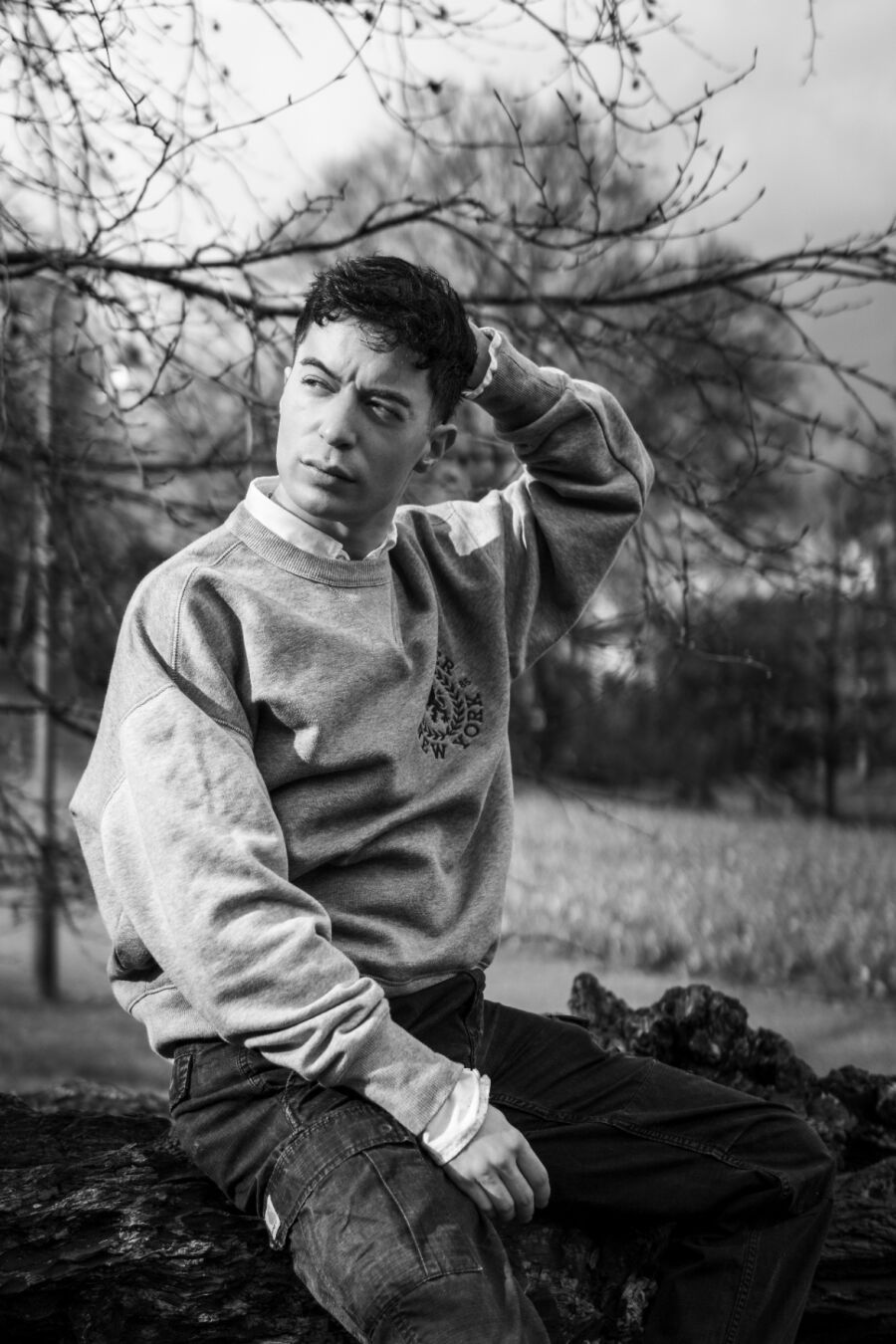 Ein schwarzweiß Foto eines männlich gelesenen Models vor einem Busch ohne Blätter. Er trägt einen grauen Pulli. Er hat eine Hand an den Hinterkopf gehoben und schaut seitlich aus dem Foto.