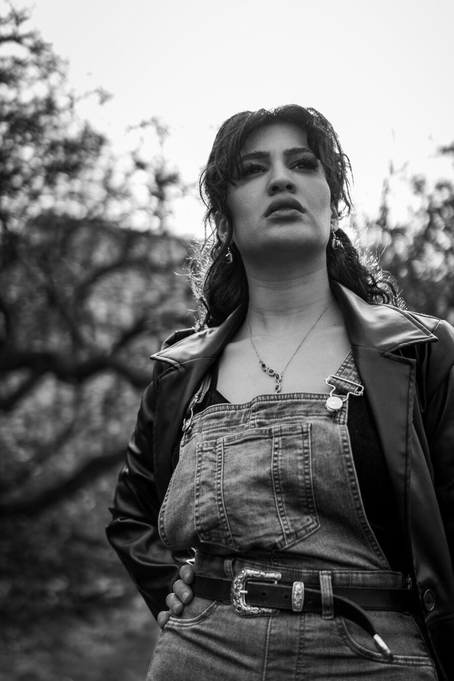 Ein schwarzweiß Foto eines weiblich gelesenen Models vor Sträuchern. Sie trägt eine Jeans-Latzhose und eine Lederjacke. Sie ist leicht von unten fotografiert und schaut über die Kamera hinweg.