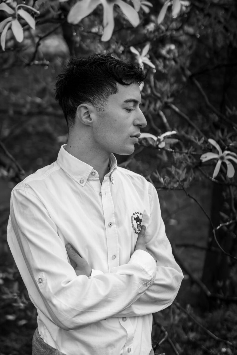 Ein schwarzweiß Foto eines männlich gelesenen Models vor einem Busch mit weißen Blüten. Er hat die Hände vor der Brust verschränkt. Sein Gesicht ist im Profil zu sehen, sein Blick ist nach unten gerichtet.