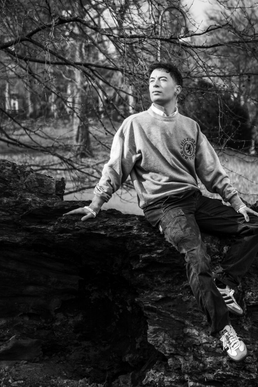 Ein schwarzweiß Foto eines männlich gelesenen Models vor einem Busch ohne Blätter. Er trägt einen grauen Pulli. Er sitzt auf einem Stein und schaut in Richtung Sonne aus dem Bild heraus.