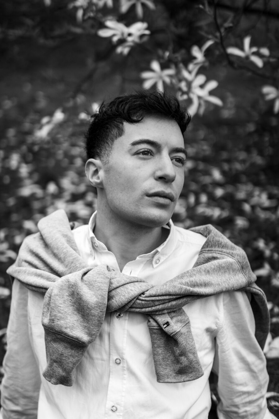 Ein schwarzweiß Foto eines männlich gelesenen Models vor einem Busch mit weißen Blüten. Er hat einen grauen Pulli um die Schultern gebunden. Er schaut verträumt aus dem Bild.