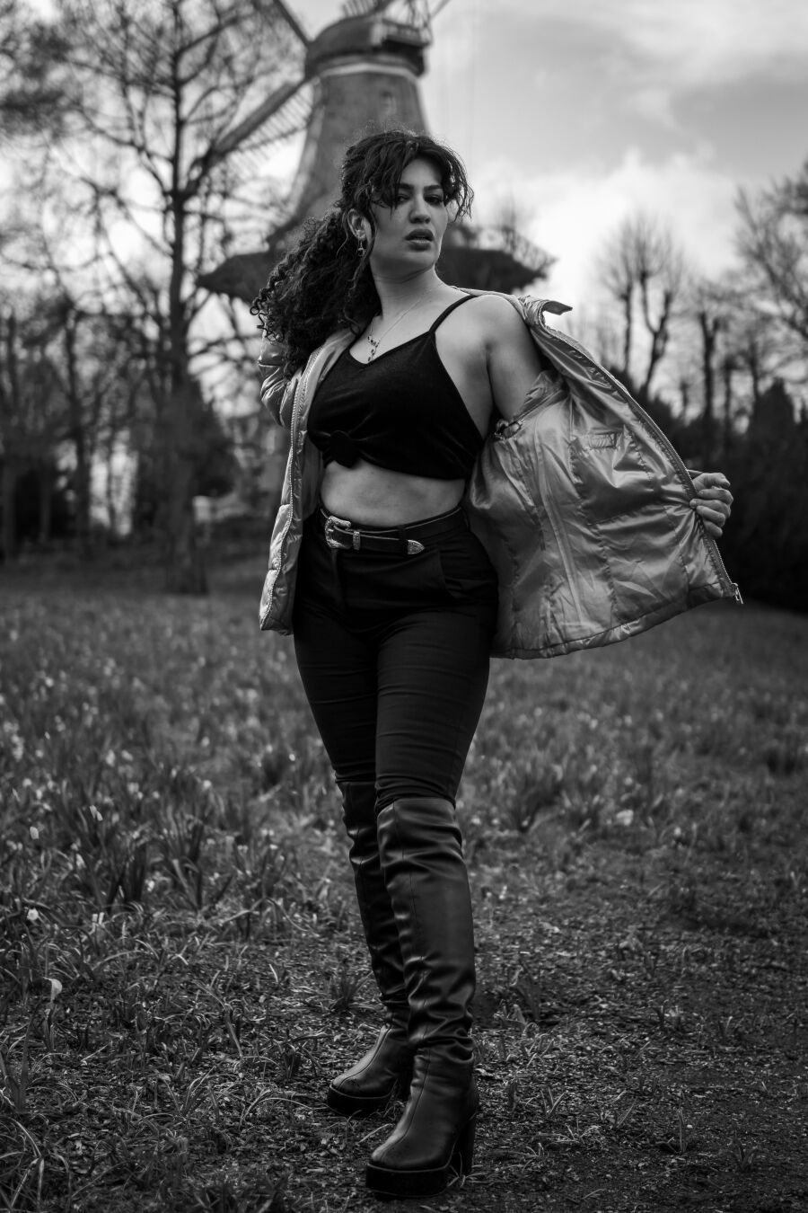 Ein schwarzweiß Foto eines weiblich gelesenen Models vor einer Windmühle. Sie trägt ein schwarzes geknotetes Tanktop und eine helle Jacke, die sie auf einer Seite geöffnet hat. Sie schaut mit leicht geöffnetem Mund direkt in die Kamera.