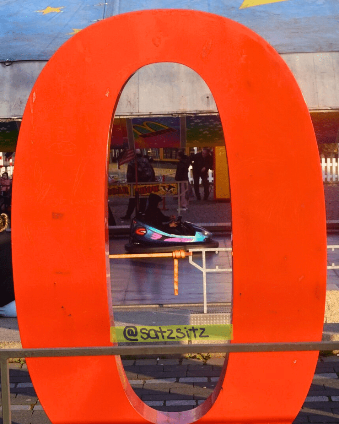 Ein großer Aufsteller der Zahl null. Durch ihren Bauch fotografiert guckt im Hintergrund ein Autoscooter durch