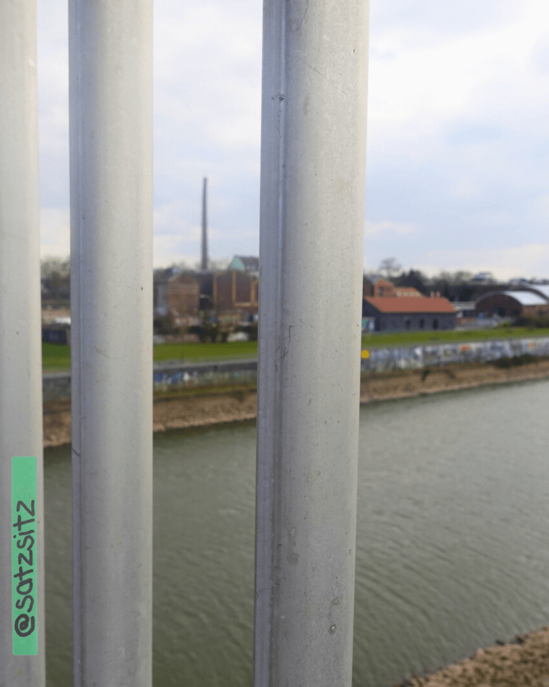 Durch geländerstäbe einer Rheinbrücke hindurch fotografiert. Der Rhein und Backsteingebäude later Industrie