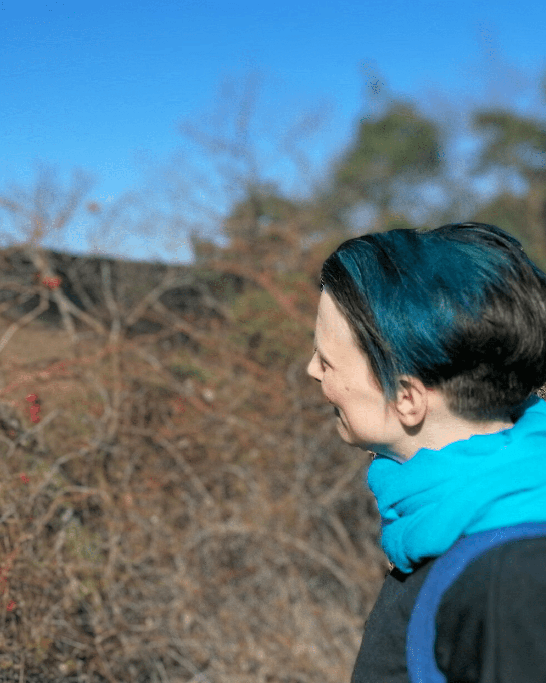 Alex vond er Seite, lacht und guckt ins Gestrüpp. Alex hat teilweise blau-türkise Haare und trägt einen hell türkisen Schal und ein dunkelgraues Oberteil