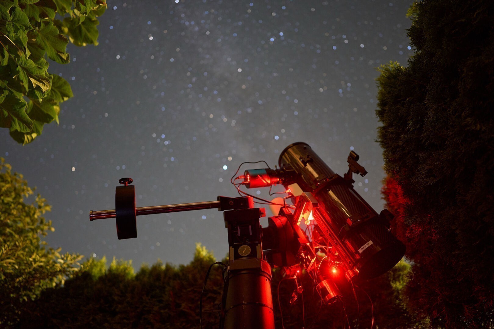Image of a backyard scene during night: A telescope mount with two different optics on top in the center of the image. Bushes and leaves are surrounding the frame of the image, with an out of focus dark sky in the background, containing the milkyway with many various stars.