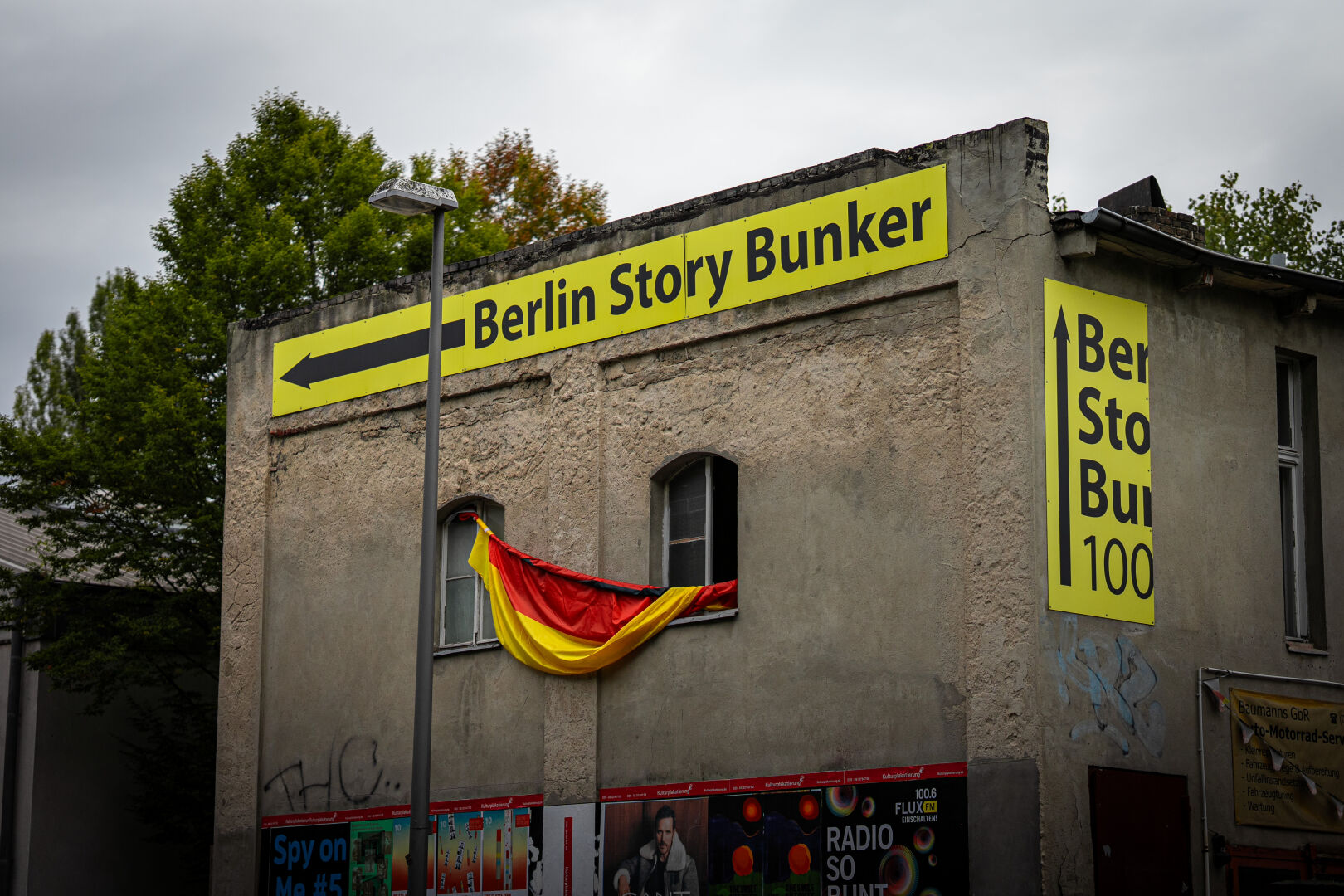Architecture shot of a concrete Building with a bright yellow sign at the top with the text "Berlin Story Bunker". One window is opened up and a Germany flag is hanging out of it.