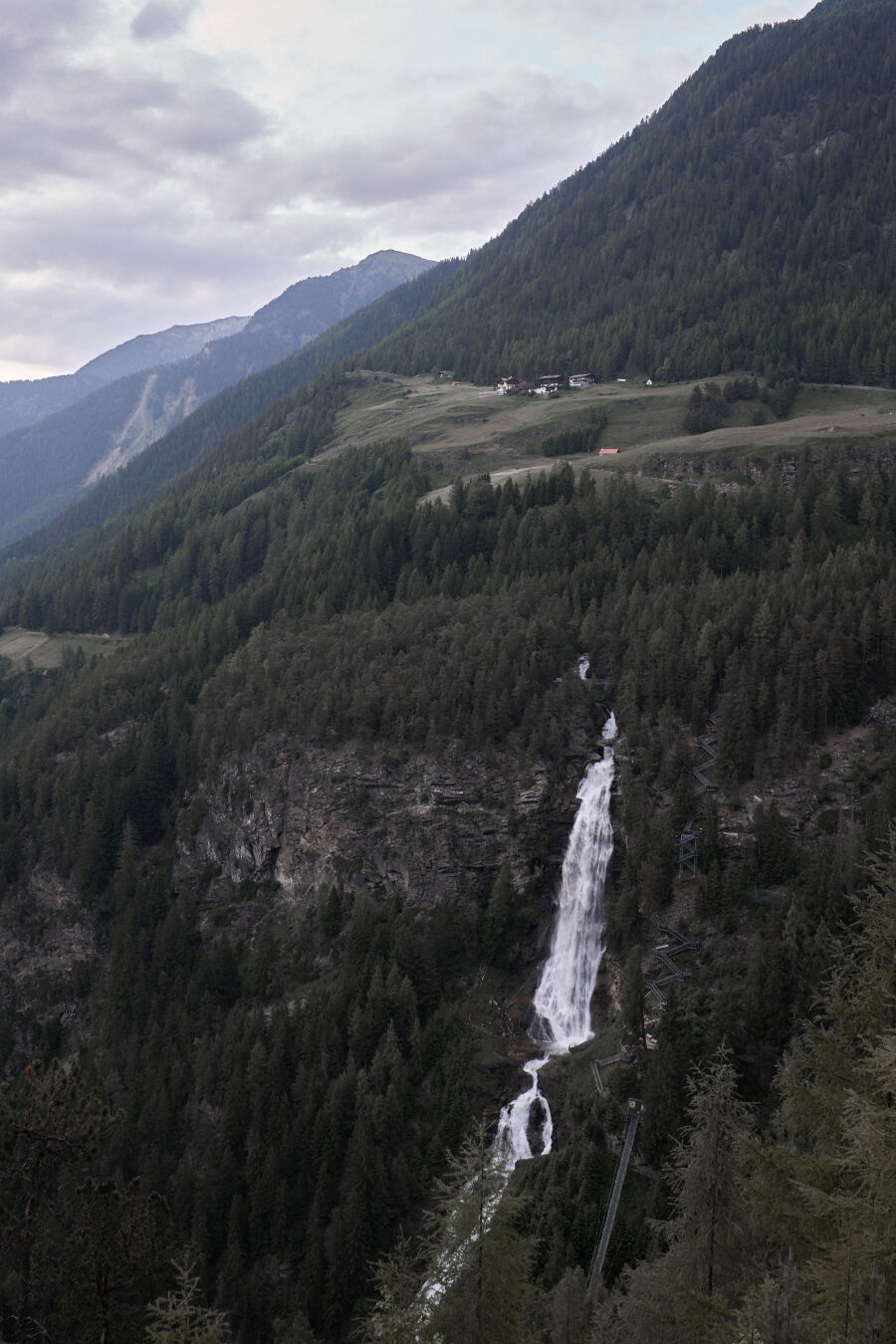 Landscape image of a long waterfall in the austrian alps. The image depics a steep mountainside with plenty of trees and a waterfall in the middle towards the lower part of the image. On top of the waterfall, there is a flat green field with a few houses. The overall mood of the image is quite dark and moody.