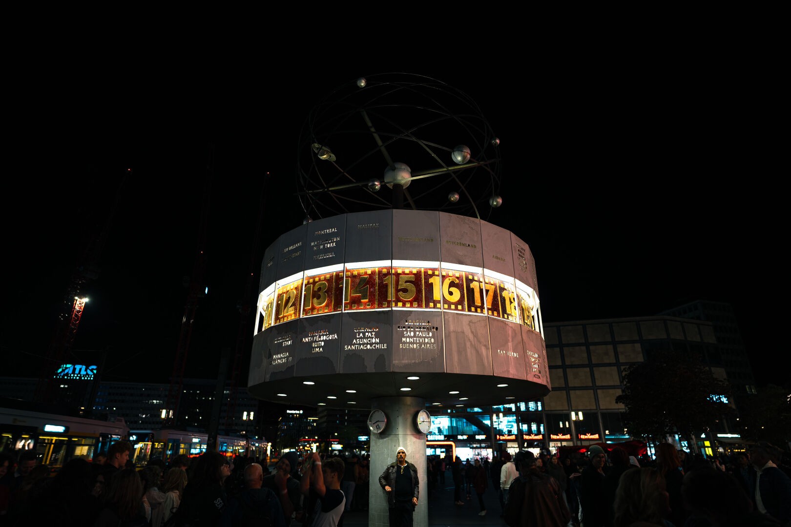 The world clock (Weltuhr) at night with a man standing right below it, being the only person brightly lit in the image. Many people around crowding the image.