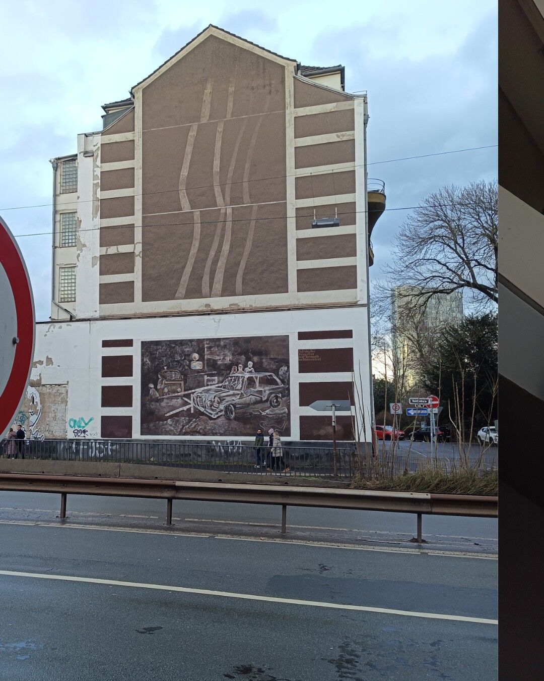 The image shows a street scene with a pedestrian forbidden sign in the foreground and a large building in the background. The building has a mural painted on its lower section, depicting a scene with people and a car. The mural is monochromatic, primarily in shades of brown. The building itself has a unique design with vertical and horizontal lines on its facade. There are a few people walking on the sidewalk in front of the building, and the street appears to be wet, suggesting recent rain. The sky is overcast, adding to the overall mood of the scene. The combination of urban elements, street art, and the weather conditions create a visually engaging and dynamic scene.